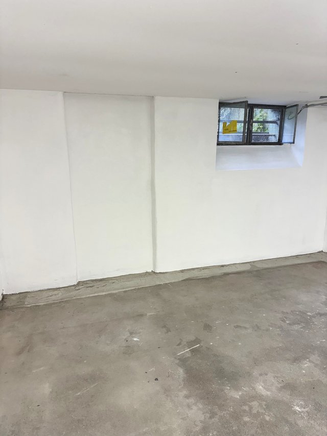Weiße Kellerwand mit kleinem vergitterten Fenster, nackter Betonboden im Vordergrund