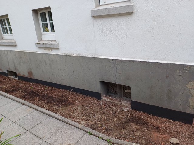 Grauer Fassadensockel mit Lüftungsgittern und freigelegtem Erdstreifen