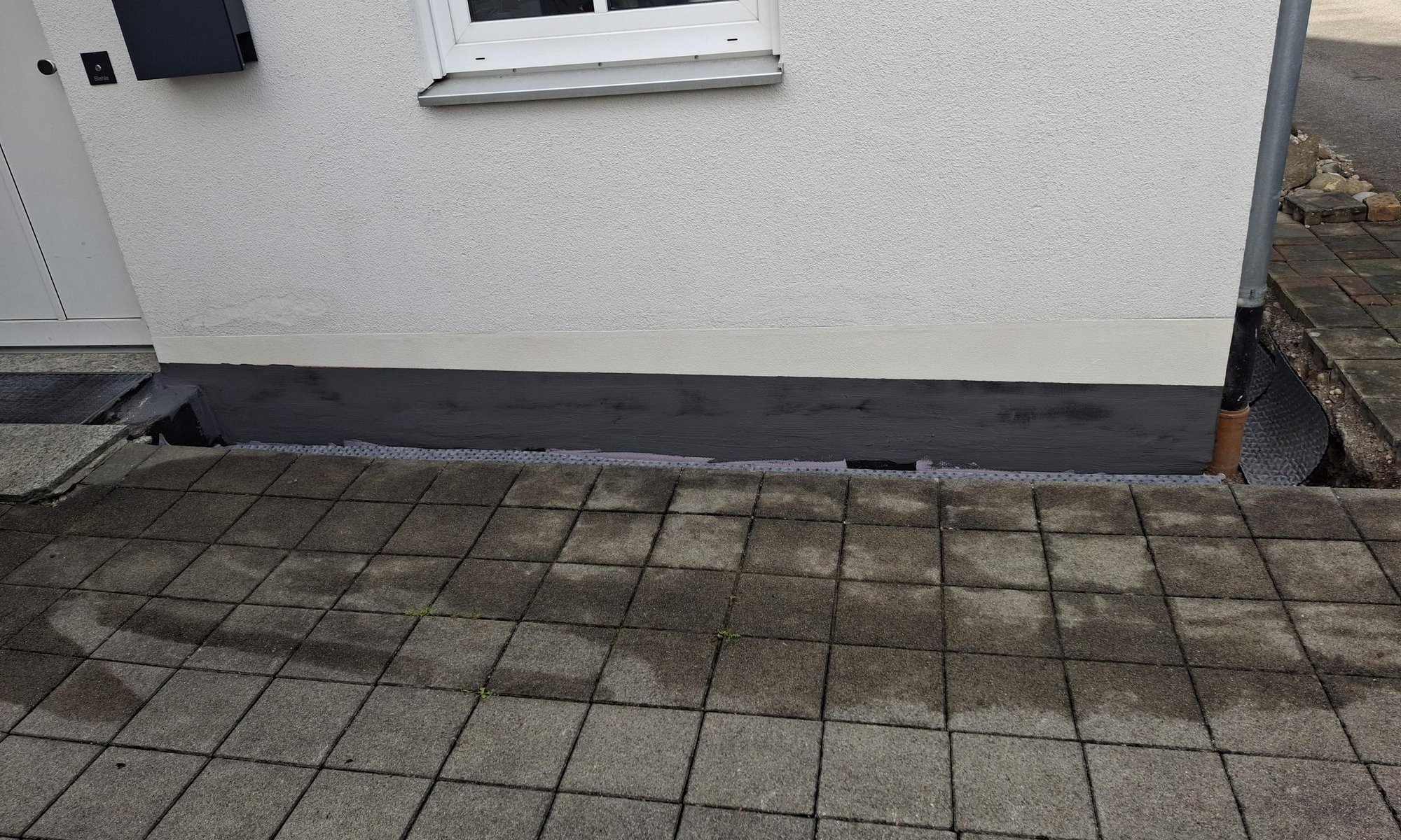 Schwarze Masse am Sockel eines Gebäudes neben gepflastertem Weg