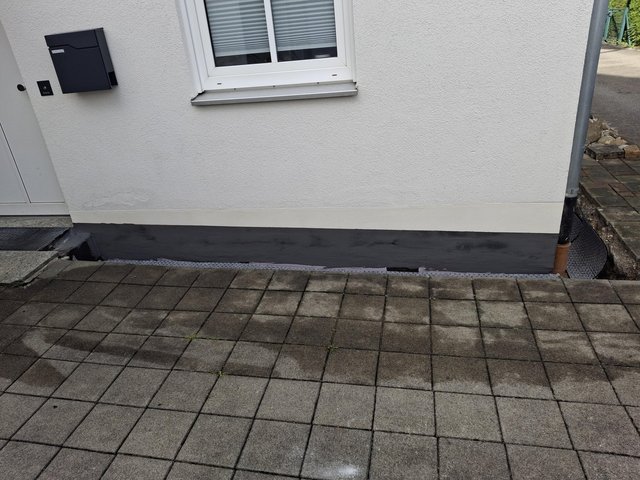 Schwarze Masse am Sockel eines Gebäudes neben gepflastertem Weg