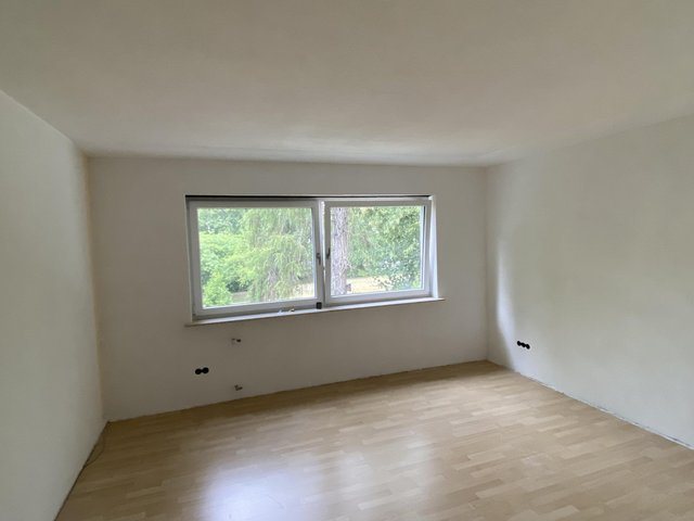 Leerer Raum mit weißer Decke, hellem Boden und breitem Fenster mit Blick ins Grüne