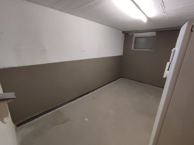 Leerer Kellerraum mit zweifarbiger Wand, kleinem Fenster und Neonleuchten an der Decke.