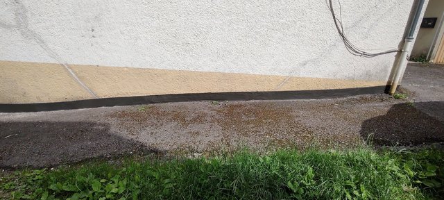 Schwarze Masse am Sockel einer verputzten Gebäudewand, davor Kiesweg und Gras