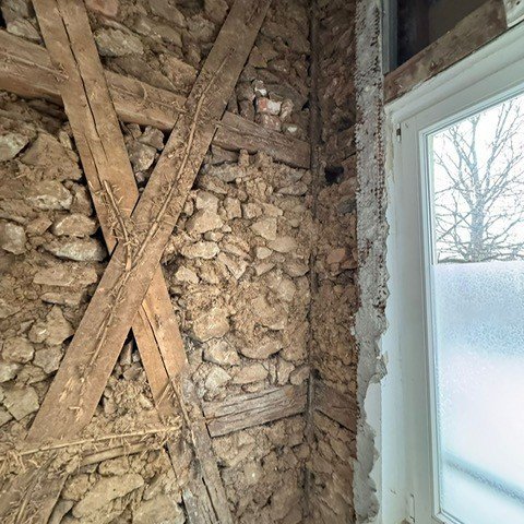 Freigelegte Steinwand mit kreuzenden Holzbalken neben Fenster.