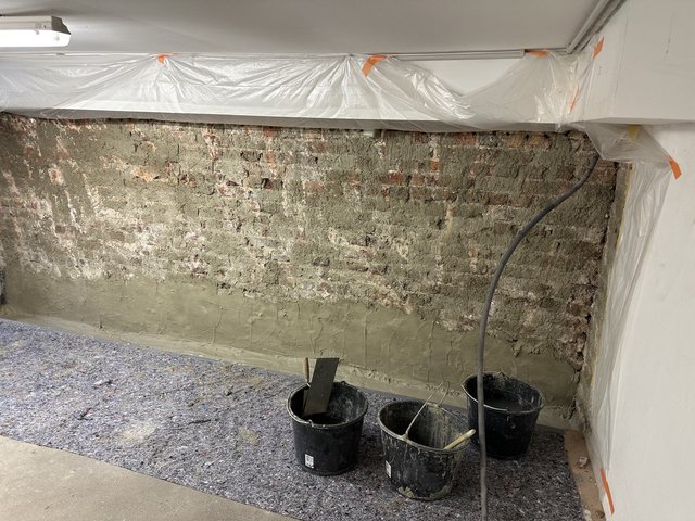 Freigelegte Kellerwand mit grauer Masse im Sockelbereich, Folienabdeckung und Eimern.