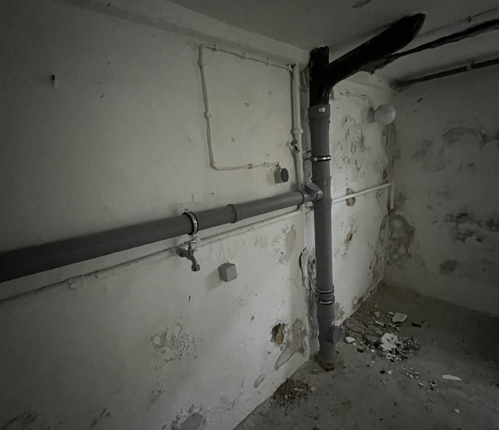 Abgeplatzter Wandputz im Kellerraum mit sichtbaren Abflussrohren und Schutt am Boden