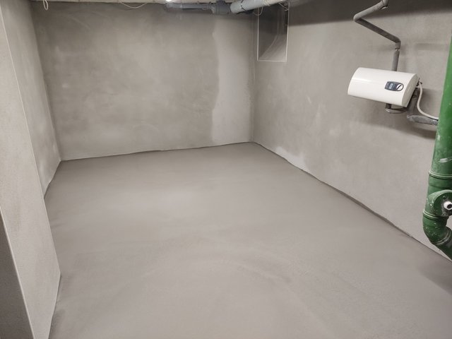 Leerer Kellerraum mit glattem grauem Boden und grauen Wänden, Rohrleitungen an der Decke.