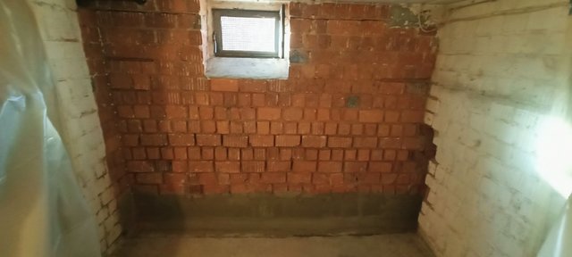 Unverputzte Ziegelwand mit kleinem Kellerfenster, dunkler Feuchterand am Sockel.