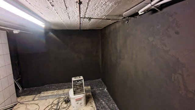 Kellerraum mit dunkel beschichteten Wänden, Deckenrohren und Baustelleneimer am Boden