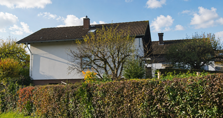 Weißes Wohnhaus mit Ziegeldach hinter Hecke und Baum, blauer Himmel mit Wolken