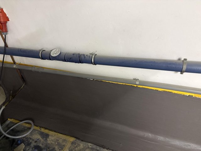 Blaues Rohr mit Reinigungsöffnung an weißer Kellerwand, darunter graue Beschichtung am Sockel.