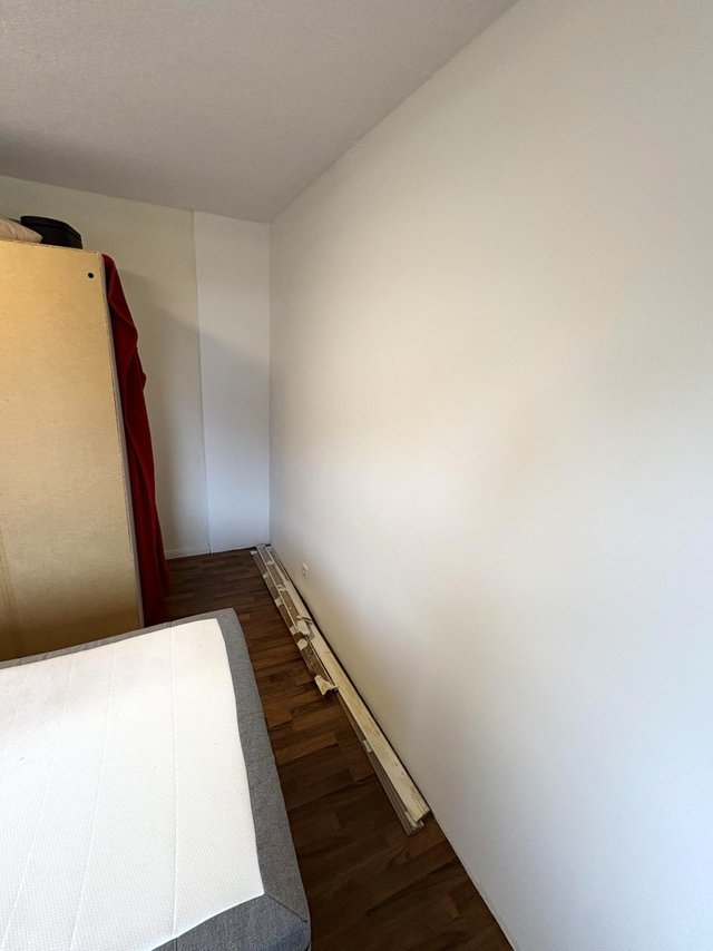 Schmale Raumecke mit weißer Wand, Bettkante und Holzleisten am Boden.