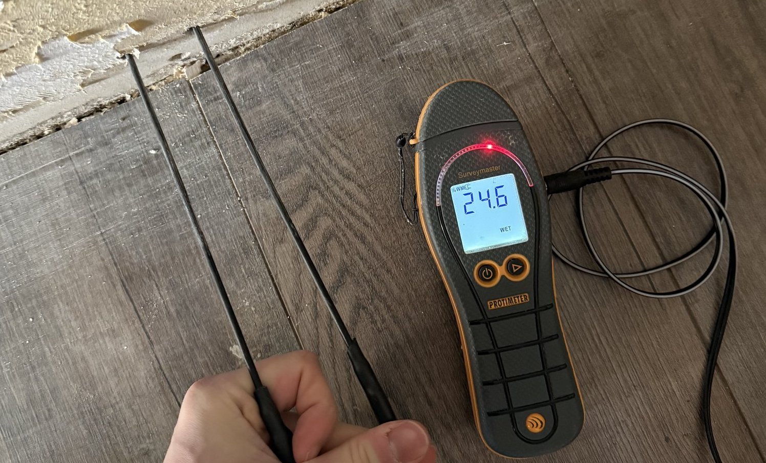 Feuchtigkeitsmessgerät misst an Bodenrand neben abblätternder Beschichtung.