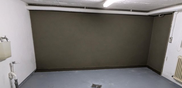 Dunkel beschichtete Kellerwand mit Rohrleitung unter der Decke und Bodenablauf.