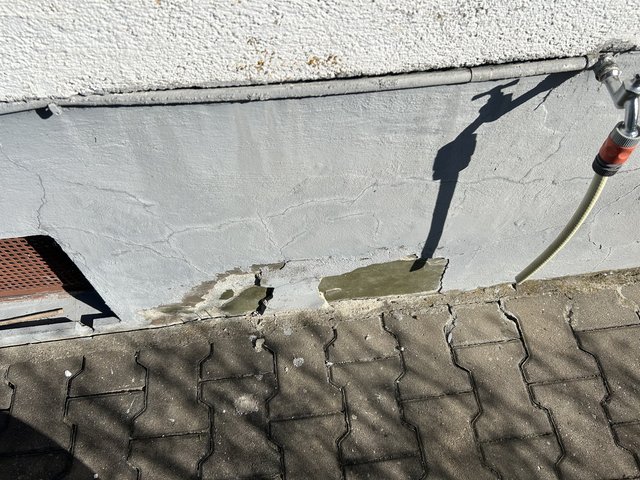 Rissiger grauer Sockelputz am Gebäude mit Ausbesserungsstellen, daneben Pflastersteine und Schlauchanschluss.
