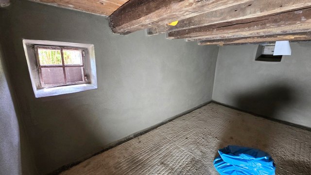 Kleiner Raum mit grauen Wänden, Holzbalkendecke, kleinem Fenster und Bodenfolie.