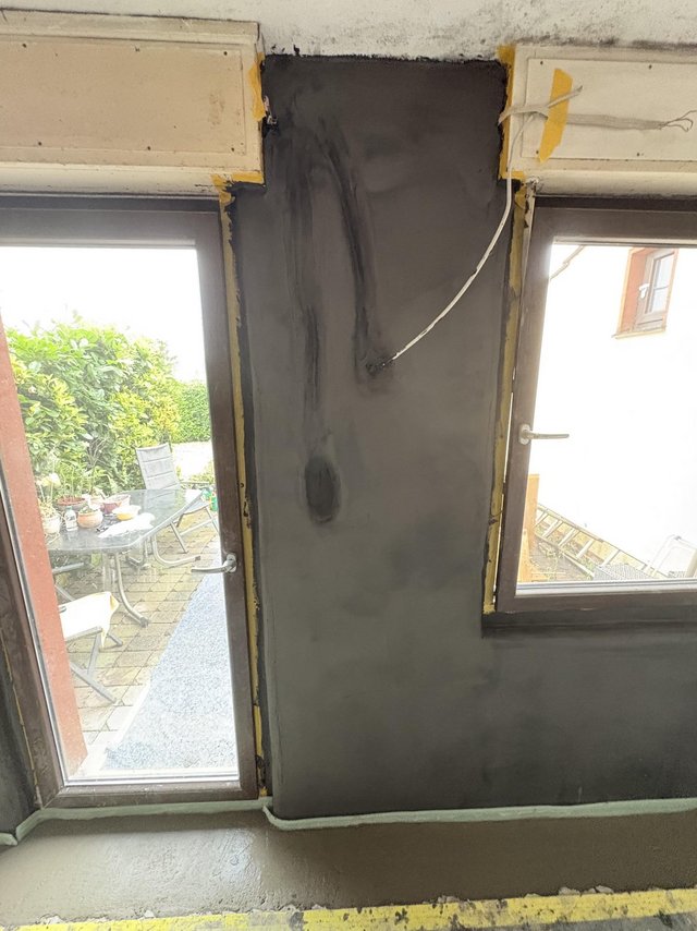 Graue Masse an Wandfläche zwischen zwei Fenstern, Kabel hängt seitlich.