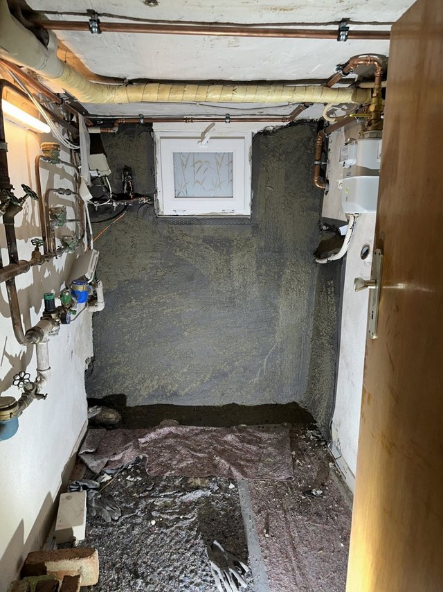 Graue Masse an Kellerwand um kleines Fenster, Technikraum mit freiliegenden Rohrleitungen