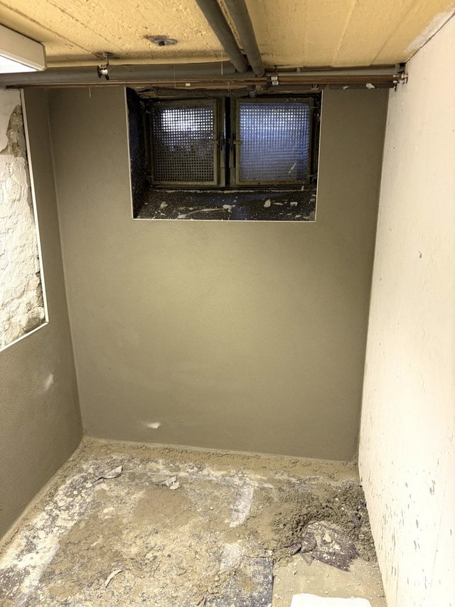 Kellerraum mit grauer Wandfläche, Fensteröffnung und Rohren an der Decke, staubiger Boden