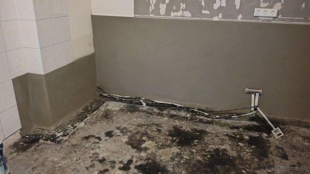 Raum mit grauer Masse an gefliester Wand und dunklen Flecken auf Betonboden