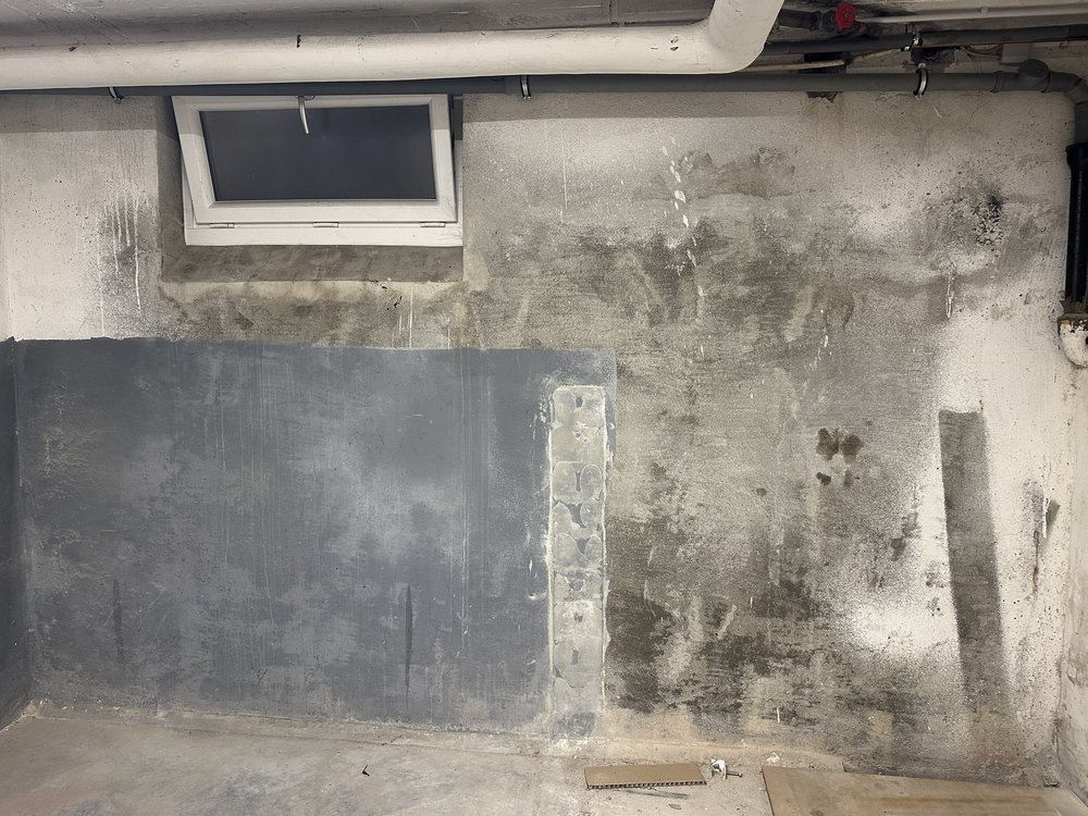 Kellerwand aus Beton mit feuchten Flecken, Rohrleitungen und kleinem Fenster.