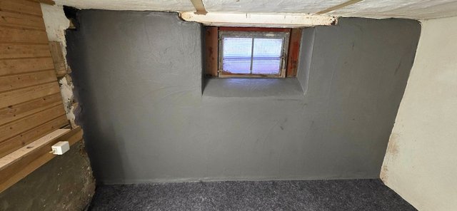 Graue Masse auf Kellerwand um kleines Fenster, naher Innenraumblick.