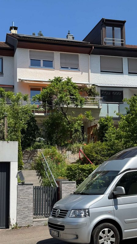 Mehrstöckiges Wohnhaus mit Balkon, Rollläden und Garten vor blauem Himmel.