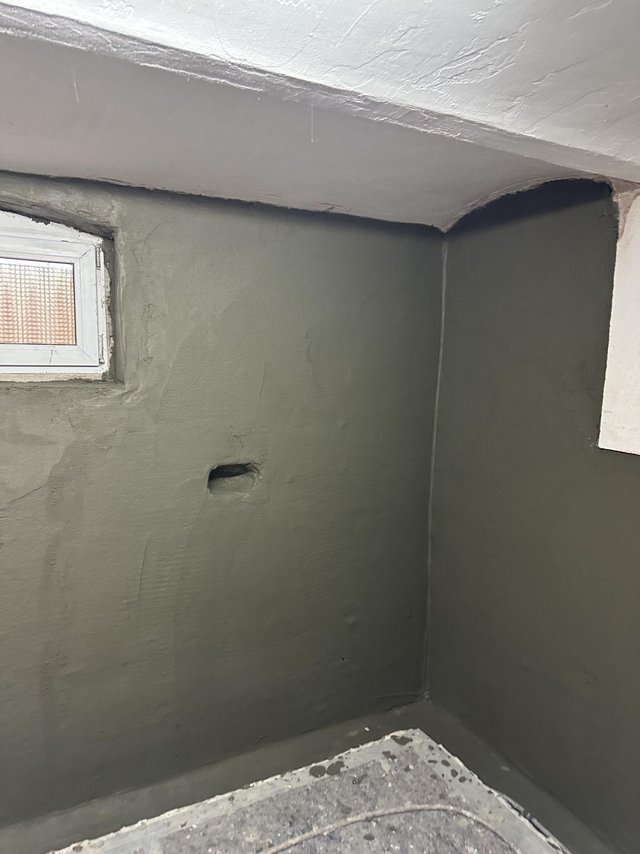Kellerwand mit frischer grauer Masse, kleines Fenster und Wanddurchführung sichtbar