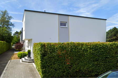 Weißes Flachdachhaus hinter hoher Hecke, seitlicher Weg und Eingang mit Treppe.