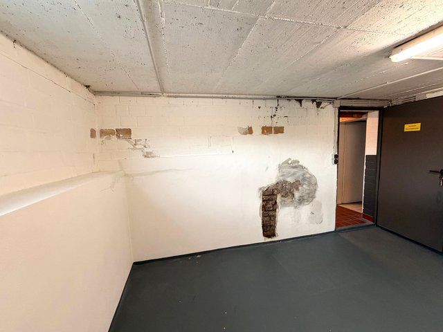 Weiße Kellerwand mit freigelegtem Mauerwerk und grauer Masse, Nahaufnahme.