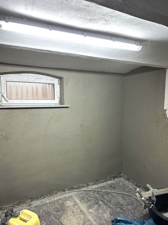 Kellerraum mit frisch verputzter Wand, Fenster und Leuchtstofflampe; Baustellenschutt am Boden