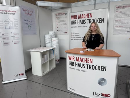 ISOTEC Mitarbeiterin steht am Messestand hinter Theke, Plakate und Eimerstapel im Hintergrund