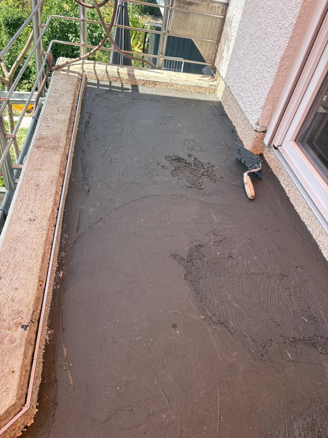 Spachtel liegt auf schwarzer Masse auf Balkonboden neben Fenster und Metallgeländer