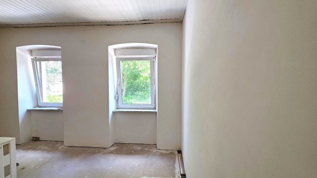 Leerer Raum mit zwei Fenstern, weiße Wände, rohe Bodenplatten und Holzdecke