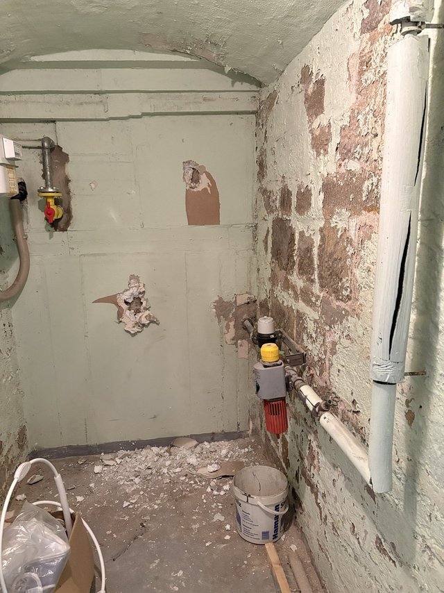 Abgeplatzter Putz und freiliegendes Mauerwerk in kleinem Kellerraum mit Rohrleitungen