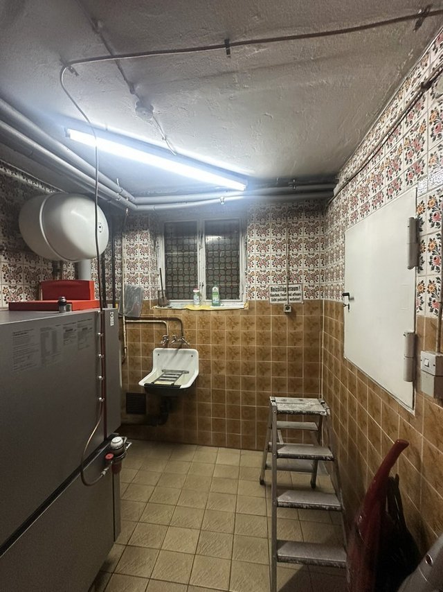 Gefliester Kellerraum mit Waschbecken, Leiter, Heizgerät und Fenster unter Neonröhre.
