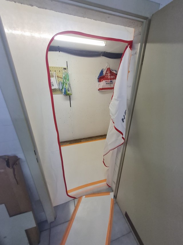 Abgeklebter Durchgang mit Reißverschlussfolie, Blick in kleinen Raum mit Lampe und Taschen