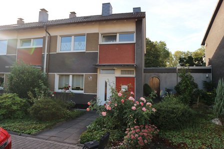 Reihenhausfassade mit weißen Fenstern und Eingangstür, davor Garten mit Rosen und Gehweg.