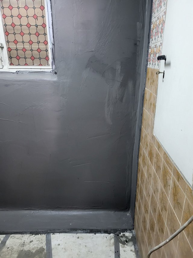 Graue Masse auf Wand und Boden neben gefliestem Bereich, Fenster links.