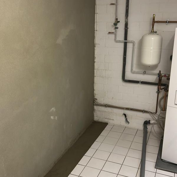 Kellerraum mit gefliesten Boden, Rohrleitungen und grauer Beschichtung an der Wand