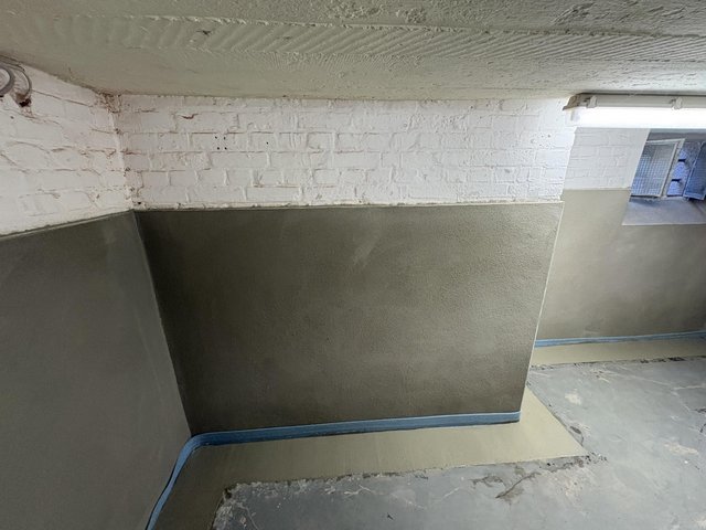 Weiße Ziegelwand über grauer Masse, Kellerraum mit Leuchte und Abklebeband am Sockel.