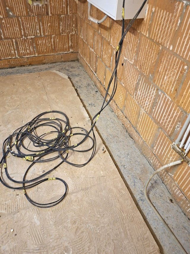 Schwarze Kabel mit gelben Etiketten liegen auf heller Estrichfläche vor Ziegelwand.