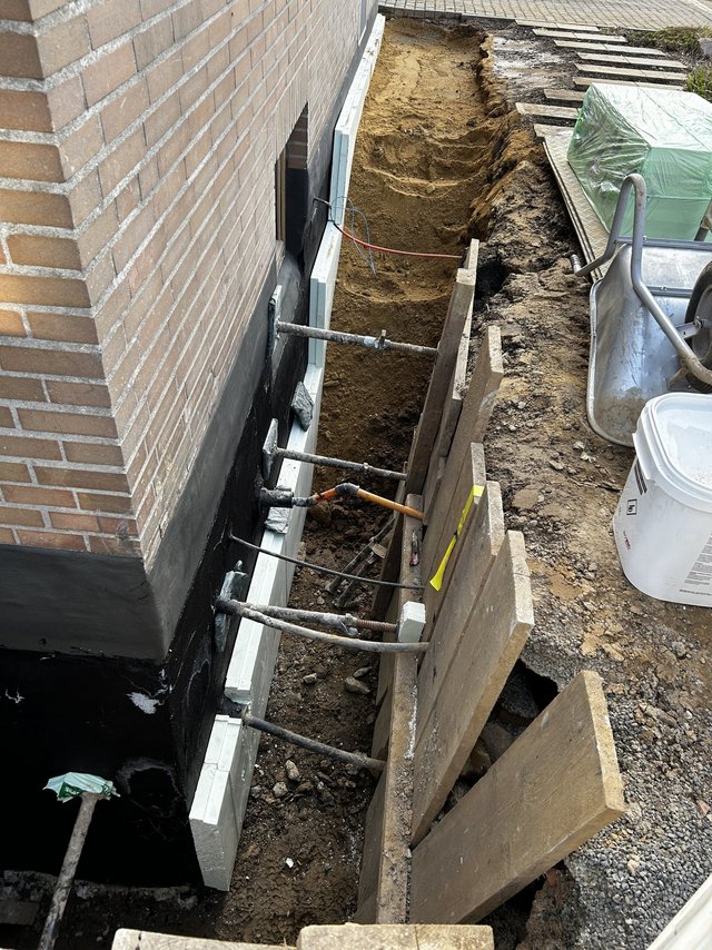 Freigelegte Kellerwand mit schwarzer Masse, heller Platte und Baugrube am Gebäude