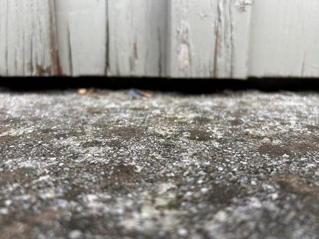Nahaufnahme von grauem Kiesbeton mit dunklen Flecken vor verwitterter, weiß gestrichener Holzwand.