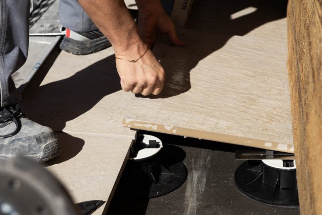 Handwerker verlegt beige Großformatplatte auf schwarzen Stelzlagern im Freien.