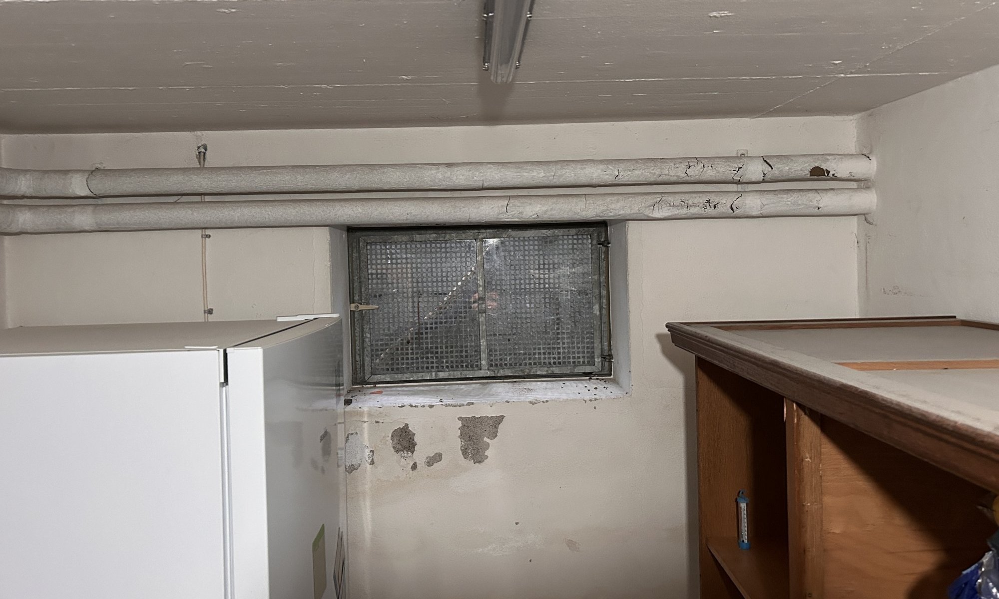 Abplatzende Beschichtung an Kellerwand unter Fenster, darüber ummantelte Rohre.