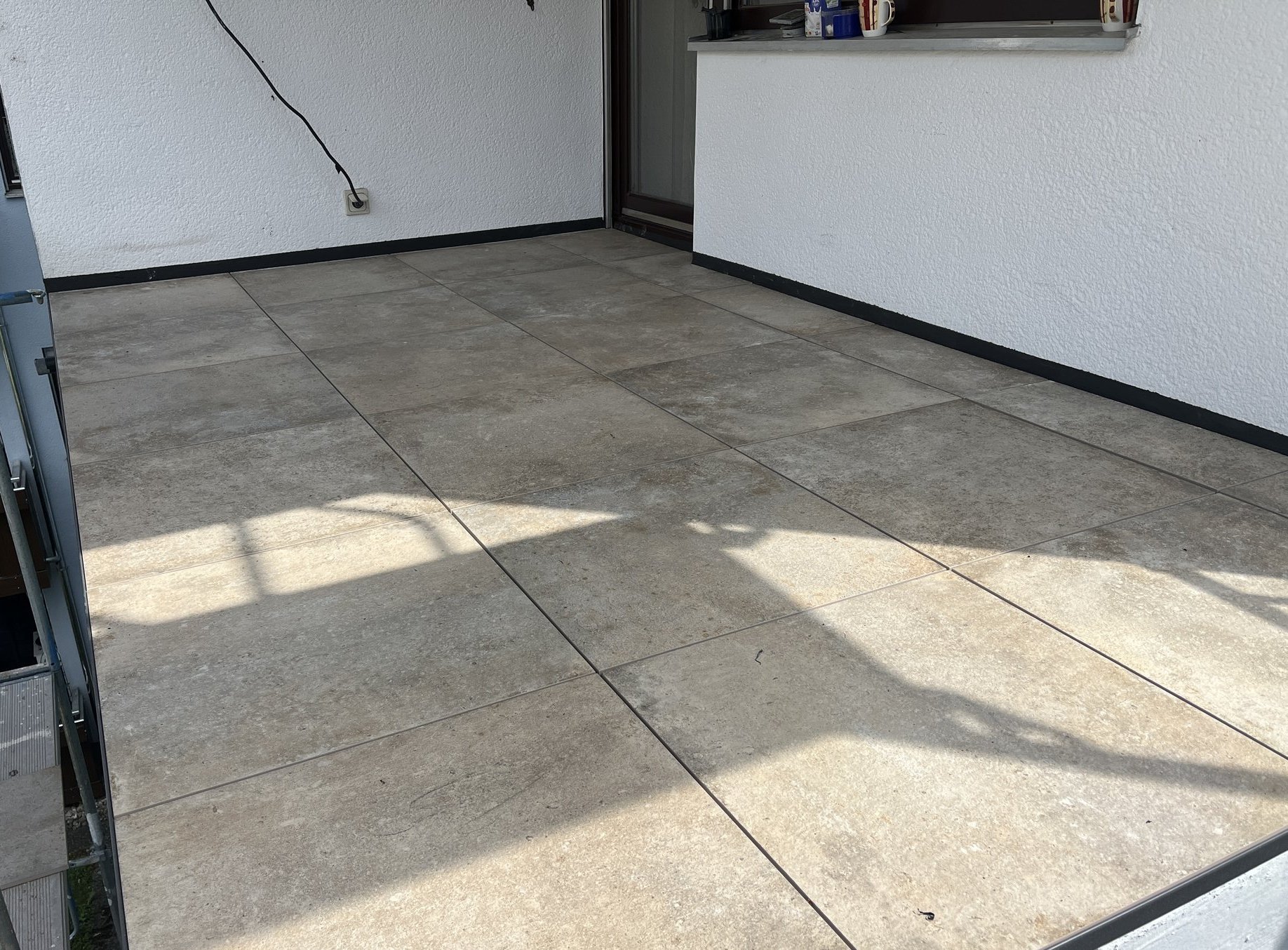 Gefliester Balkonboden mit grauen Platten vor weißer Fassade und Schiebetuer.