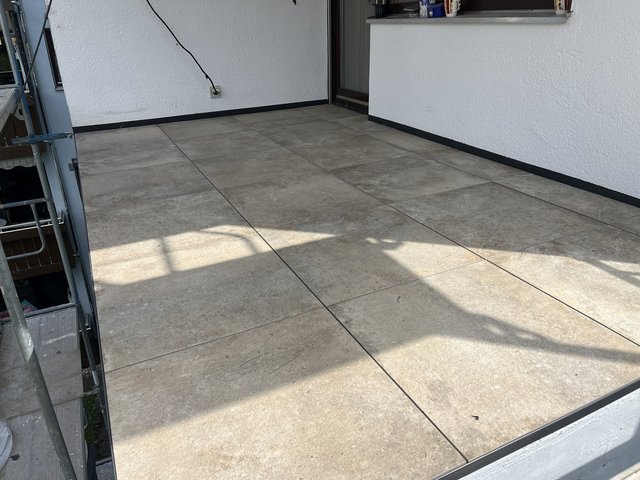 Gefliester Balkonboden mit grauen Platten vor weißer Fassade und Schiebetuer.