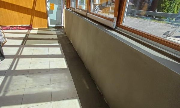 Sonniger Wintergarten mit Holzrahmenfenstern, Fliesenboden und grauer Masse entlang der unteren Fensterbrüstung