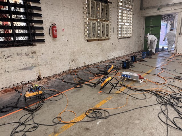 Zwei Arbeiter verlegen Kabel und Geräte entlang einer gemauerten Kellerwand.